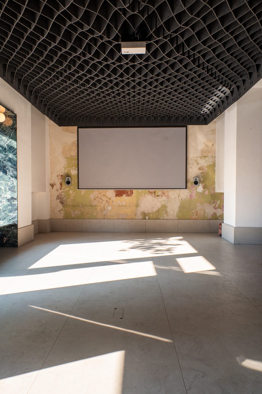 Die heruntergelassene Leinwand vor der patinierten Wand, links eine grün geäderte Marmorlaibung, Sonnenlicht in Flecken auf dem Boden.