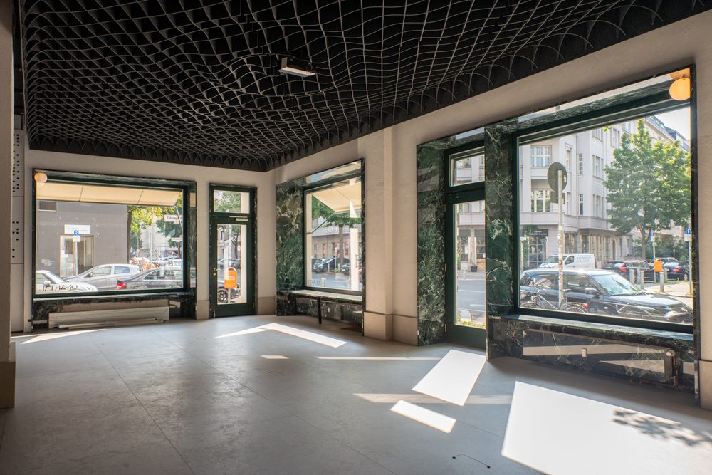 Der Saal: dunkle Rasterdecke, grün geäderte Marmorlaibungen an der Fensterfront, Sonnenlicht in Streifen auf dem Boden, dahinter die Straße.