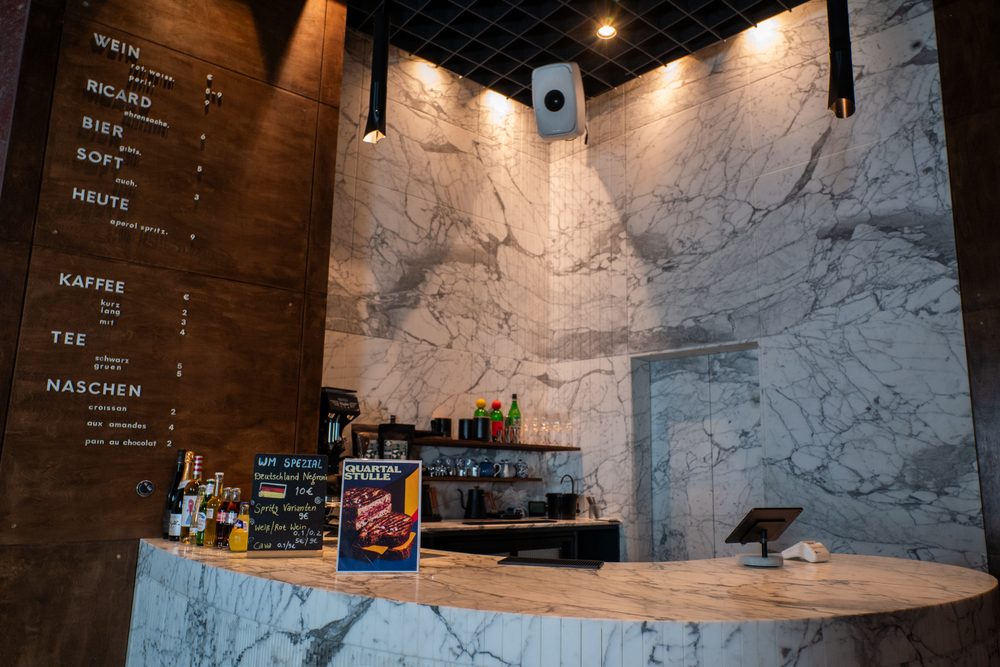 Der Tresen aus weißem Marmor vor der geäderten Wand, links die Holztafel mit WEIN, KAFFEE, TEE und NASCHEN, darauf Flaschen und die Maschine.